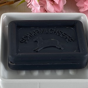 Könnte beinhalten: Ein dunkelgraues Seifenstück mit der Prägung "SCHAFMILCHSEIFE" und einer Schafillustration, liegt in einer weißen Keramikseifenschale. Rosa Blüten sind im Hintergrund zu sehen.