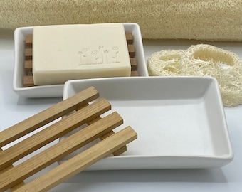 May include: Two white ceramic soap dishes, one with a wooden slatted base and the other with a bar of soap. The soap has a floral design embossed on it.