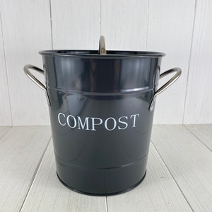 Könnte beinhalten: Ein grauer Komposteimer aus Metall mit einem silbernen Griff und dem Wort "COMPOST" in weiß auf der Vorderseite.