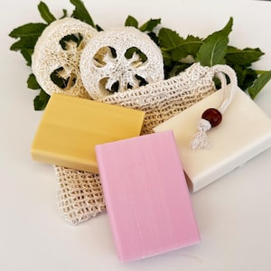 Könnte beinhalten: Drei Seifenstücke, eines gelb, eines rosa und eines weiß, liegen auf einer weißen Oberfläche. Eine kleine, natürliche, gewebte Tasche mit Kordelzug ist mit zwei Luffaschwämmchen gefüllt.