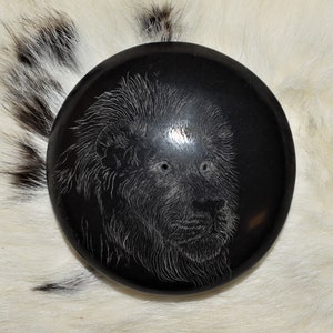 Puede incluir: Un objeto decorativo redondo y negro con una imagen grabada detallada de la cara de un león. La melena y los rasgos faciales del león están intrincadamente tallados. El objeto está sobre un fondo de piel blanca y negra.