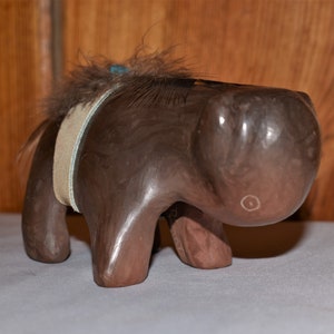 May include: A small, brown, carved animal figurine with a feathered mane and a white leather strap around its body.