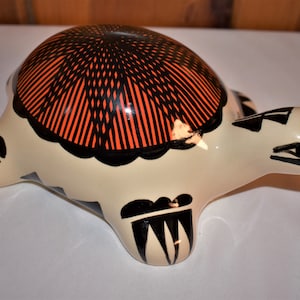 May include: A ceramic turtle figurine with a black, orange, and white geometric design. The turtle is painted with black lines and orange stripes on its shell.