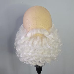 Puede incluir: Un conjunto de peluca y barba de Papá Noel. La peluca es de color beige claro, y la barba es blanca y voluminosa. La peluca y la barba están montadas sobre un soporte negro sobre un fondo gris claro. Ideal para eventos navideños.