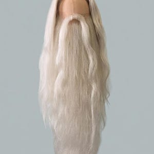 Op de afbeelding: Een lange, witte pruik van synthetisch haar met een volle, vloeiende baard en snor bevestigd aan een kanten kapbasis. Het haar is gestyled met een middenscheiding en hangt recht naar beneden in lange, getextureerde lokken voor volume.