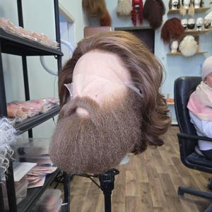 May include: A brown wig with a matching beard and mustache, mounted on a mannequin head. The wig features long, wavy hair and a full beard. Other wigs are visible on shelves in the background.