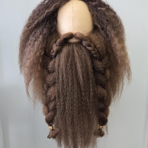 Op de afbeelding: Een bruine pruik met een lange, gevlochten baard. De pruik heeft een lichtbruin hoofd en donkerbruin, krullend haar. De baard is donkerder bruin en is gestyled in twee vlechten, elk versierd met een gouden kraal. De pruik is gemonteerd op een mannequinhoofd.