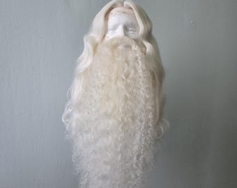 May include: A white wig and beard set displayed on a mannequin head. The wig features long, wavy hair, and the beard is a full, curly style. The set is likely for costume or theatrical use, with a focus on a classic, aged appearance.