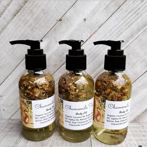 May include: Three amber glass bottles with black pump tops filled with a yellow liquid and dried chamomile flowers. The bottles have white labels with the text "Chamomile Body Oil" and a list of ingredients.