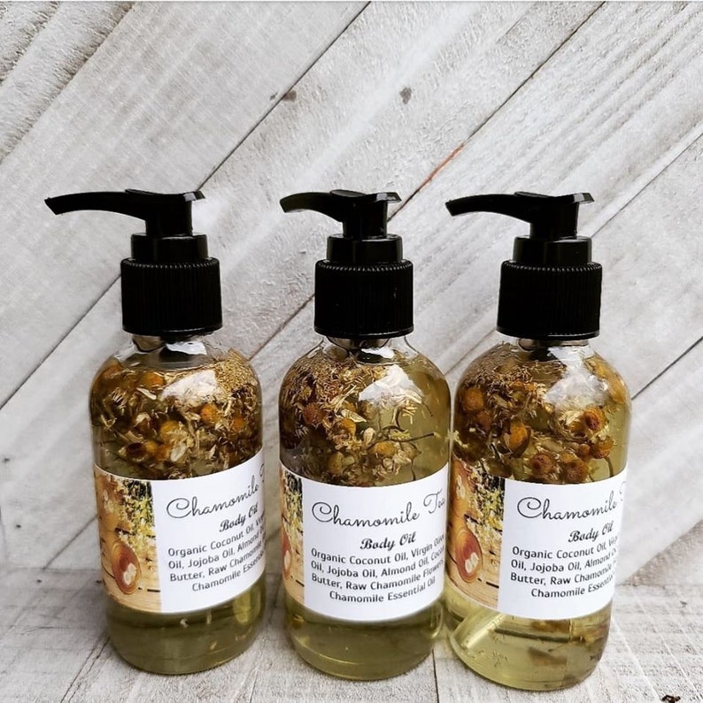 May include: Three amber glass bottles with black pump tops filled with a yellow liquid and dried chamomile flowers. The bottles have white labels with the text "Chamomile Body Oil" and a list of ingredients.