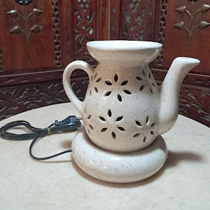 May include: White ceramic electric oil diffuser shaped like a teapot with a floral design. The diffuser has a cord and plug.