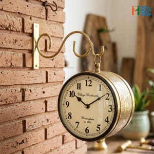 May include: A double-sided wall-mounted clock with a brass finish, featuring a cream-colored face with black Arabic numerals and the text Village Clockworks Birmingham est. 1782. The clock hangs from a decorative curved brass metal wall bracket.