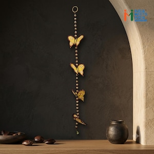 Puede incluir: Una decoración de pared vertical que consiste en una cuerda con pequeñas cuentas de madera redondas intercaladas con cuatro adornos de mariposas amarillas. Las mariposas tienen bordes marrón oscuro y patrones detallados. Incluye un anillo de metal en la parte superior.