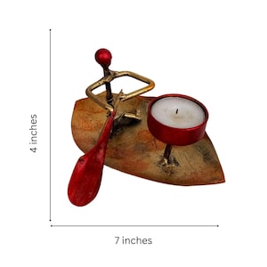 May include: A red metal candle holder shaped like a person rowing a boat. The boat is on a gold leaf-shaped base. The candle holder is 7 inches wide and 4 inches tall.