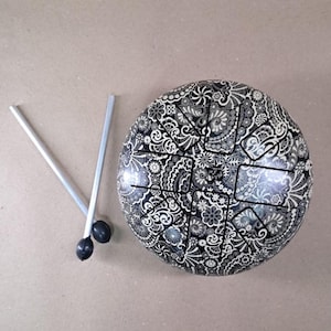 May include: A steel tongue drum with a black and white floral pattern. Two silver drum mallets with black tips are positioned to the left of the drum. The drum has a series of cut-out tongues that produce different tones when struck.