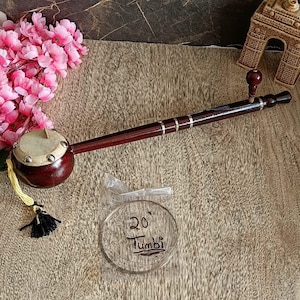May include: A brown wooden Tumbi with a round, brown, leather-covered drum head. The Tumbi has a silver string and a small, brown knob at the end. The Tumbi is on a wooden surface with a small, clear plastic bag containing a silver string labelled "20' Tumbi".