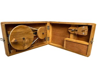May include: A wooden spinning wheel in a wooden box. The spinning wheel has a large wooden wheel and a smaller wooden wheel. The box is open and the spinning wheel is visible.