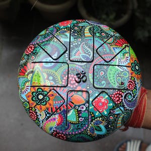 Könnte beinhalten: Ein schwarzes Stahl-Handpan mit einem farbenfrohen Paisley-Muster. Das Handpan hat acht Töne und ist mit einem Om-Symbol verziert.