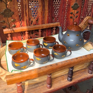 Op de afbeelding: Een set van zes zwarte en bruine keramische theekopjes en een bijpassende theepot met een gesneden houten dienblad. De kopjes en de theepot hebben een rustiek, handgeschilderd ontwerp.