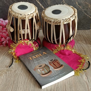 May include: Two brown tabla drums with white tops and leather straps. The drums are sitting on pink and gold fabric cushions. A book titled "Tabla: Theory and Practice of Tabla" by Sadanand Naimpally is in the foreground. The book has a black cover with white text and a photo of two tabla drums.