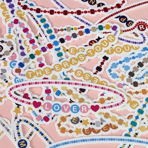 May include: A collection of colourful beaded necklaces with various words and phrases, including "Speak Now", "Lover", "The Eras", "Folklore", "Red", "1989", "Midnights", and "Reputation".