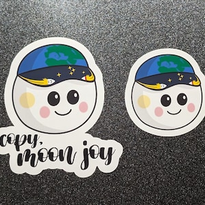 May include: Two white stickers with a cartoon character wearing a blue and green Earth hat with stars. The character has a smiling face with rosy cheeks. One sticker has the text "copy. moon joy" in black script.