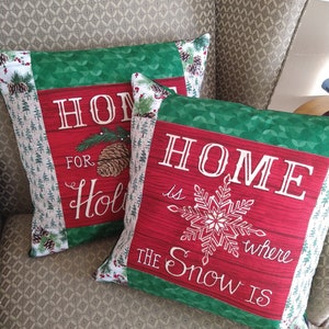 May include: Two decorative Christmas pillows with red and green fabric. One pillow reads "HOME for Holidays" and the other reads "HOME is where the Snow IS". The pillows have a festive design with pinecones and snowflakes.