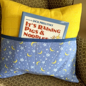 May include: A yellow pillow with a blue pocket, holding a book titled "It's Raining Pigs & Noodles." The pocket is patterned with white stars and yellow crescent moons. The pillow is on a patterned chair.