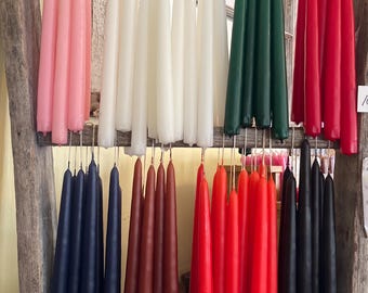 May include: A collection of long, slender taper candles hanging in rows from a wooden rack. The candles are available in a variety of solid colors, including pink, white, dark green, bright red, navy blue, deep brown, orange, and black.