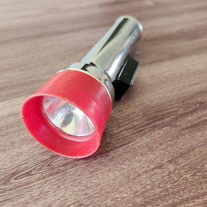 May include: A silver flashlight with a red bezel and a black clip. The flashlight is lying on a wooden surface. The light is on.