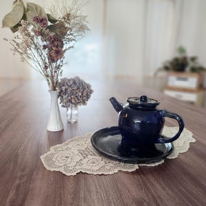 May include: A navy blue ceramic teapot with a matching saucer sits on a cream-colored lace doily. A white vase with dried flowers and a small glass vase with a purple flower are also on the wooden table.