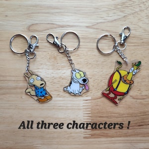 May include: Three cartoon character keychains with silver key rings and clasps. The characters are a yellow kangaroo, a white dog with a pink tongue, and a yellow and red character. The text "All three characters!" is below the keychains.