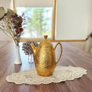 May include: A gold-colored teapot with a textured surface sits on a cream-colored lace doily. The teapot has a lid and a curved handle. Dried flowers in a white vase and a window are in the background. The table is made of wood.