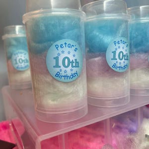 May include: Clear plastic containers filled with cotton candy in layers of blue, white, and pink. Each container has a blue sticker that reads "Petar's 10th Birthday". The containers are arranged on a clear, pink display shelf.