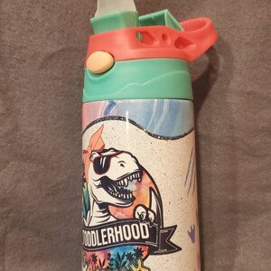 Puede incluir: Una botella de agua colorida con un dise&ntilde;o de dinosaurio. La botella tiene una tapa y un asa de color turquesa, coral y blanco. El cuerpo de la botella presenta un dinosaurio de dibujos animados con gafas de sol, con las palabras "Toddlerhood Walk the Park".
