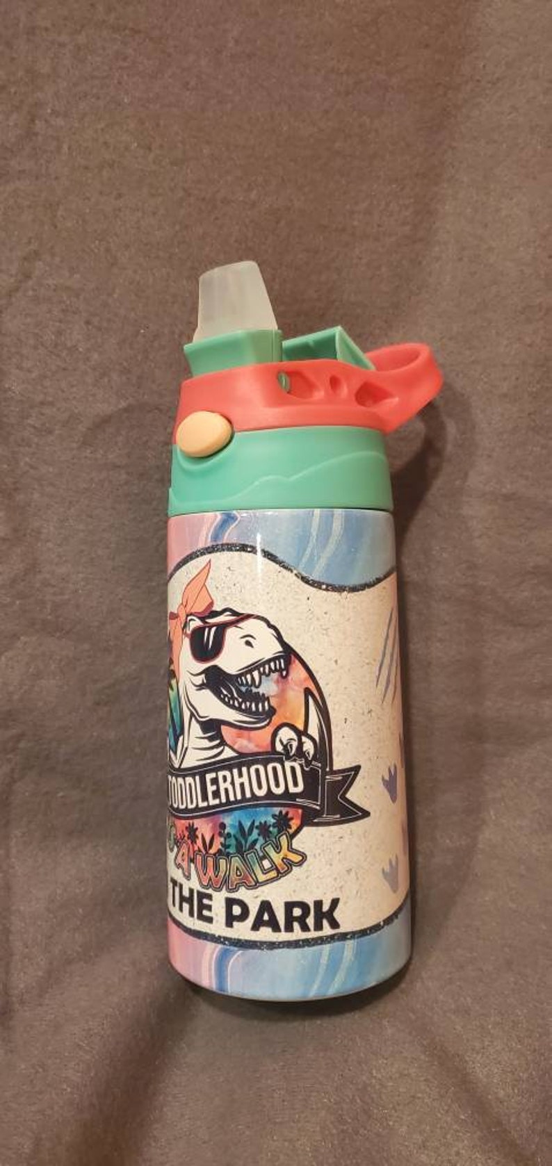 Puede incluir: Una botella de agua colorida con un dise&ntilde;o de dinosaurio. La botella tiene una tapa y un asa de color turquesa, coral y blanco. El cuerpo de la botella presenta un dinosaurio de dibujos animados con gafas de sol, con las palabras "Toddlerhood Walk the Park".