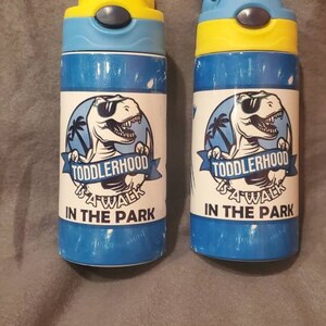 Puede incluir: Dos botellas de agua azules y amarillas con un dise&ntilde;o de dinosaurio y el texto "TODDLERHOOD A WALK IN THE PARK". Las botellas tienen una tapa abatible y est&aacute;n dise&ntilde;adas para ni&ntilde;os.