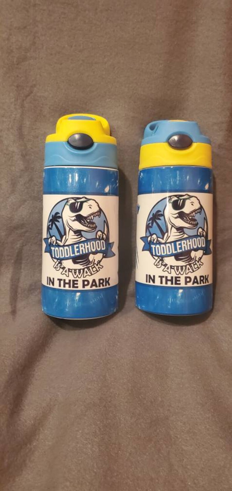Puede incluir: Dos botellas de agua azules y amarillas con un dise&ntilde;o de dinosaurio y el texto "TODDLERHOOD A WALK IN THE PARK". Las botellas tienen una tapa abatible y est&aacute;n dise&ntilde;adas para ni&ntilde;os.