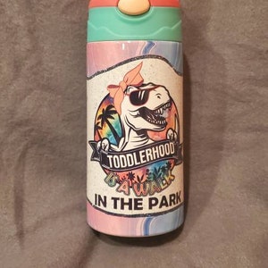 Puede incluir: Una botella de agua colorida con un dise&ntilde;o de dinosaurio con gafas de sol y un lazo. La botella tiene un esquema de color rosa, turquesa y blanco, con el texto "Toddlerhood Squad In The Park".