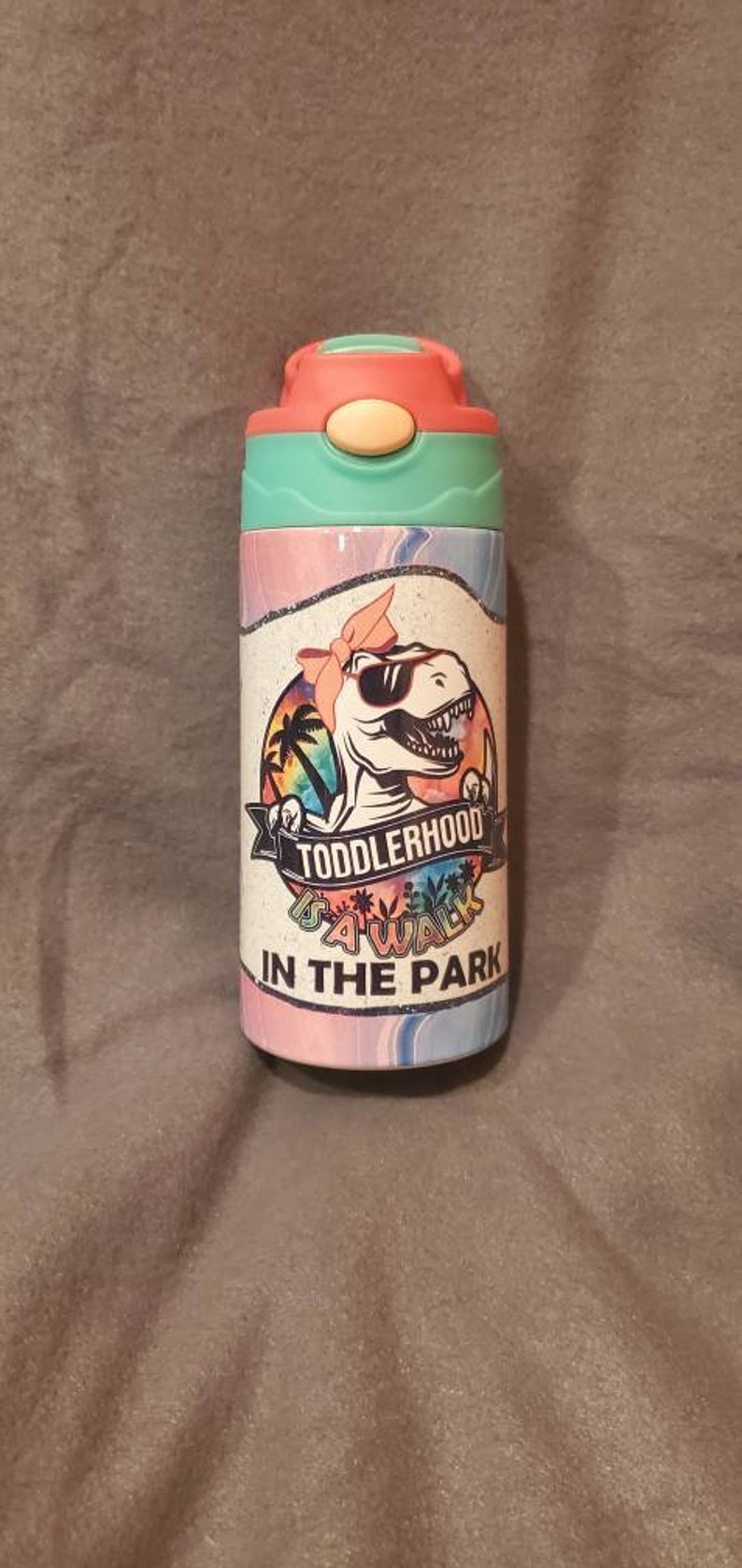 Puede incluir: Una botella de agua colorida con un dise&ntilde;o de dinosaurio con gafas de sol y un lazo. La botella tiene un esquema de color rosa, turquesa y blanco, con el texto "Toddlerhood Squad In The Park".
