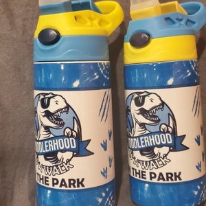 Puede incluir: Dos botellas de agua azules y blancas con tapas amarillas y azules. Cada botella presenta un gr&aacute;fico de dinosaurio y el texto "Toddlerhood Walk the Park".
