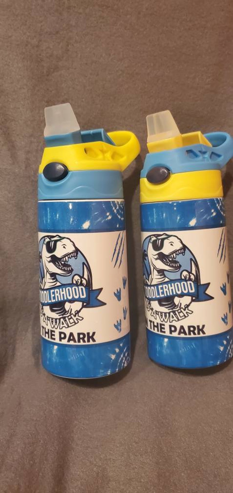 Puede incluir: Dos botellas de agua azules y blancas con tapas amarillas y azules. Cada botella presenta un gr&aacute;fico de dinosaurio y el texto "Toddlerhood Walk the Park".