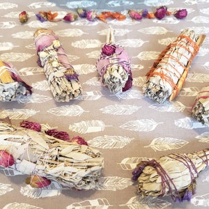 Könnte beinhalten: Eine Auswahl an Salbei-Bündeln, verziert mit getrockneten Blumen und Bindfäden. Die Bündel sind mit verschiedenfarbigen Bindfäden umwickelt und mit rosa, orangefarbenen und lila getrockneten Blumen verziert. Die Salbei-Bündel sind auf einem grauen Stoff mit weißem Federmuster angeordnet.