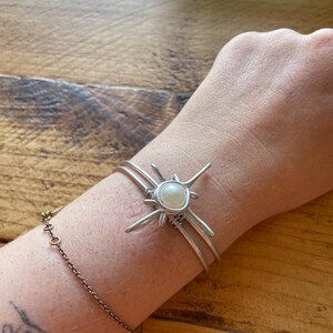 May include: A silver-toned wire cuff bracelet featuring a central white pearl-like bead. The wire is wrapped around the bead in a starburst pattern with four radiating points. The bracelet consists of two parallel silver-toned wire bands.