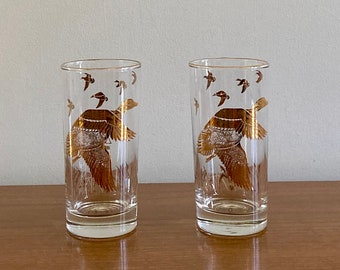 May include: Two clear glass drinking glasses with a gold design of flying ducks. The glasses are on a wooden surface.