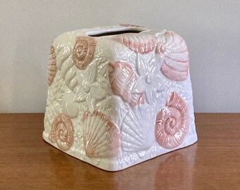 May include: A square, white ceramic tissue box with a raised seashell design in pink. The tissue box is on a wooden surface, evoking a coastal theme. The dimensions are approximately 15 cm x 15 cm.