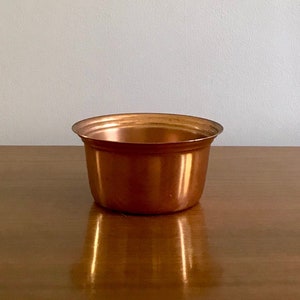 May include: A small, copper-coloured planter pot with a slightly flared rim. The pot is sitting on a wooden surface.