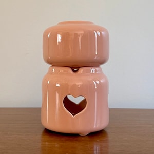 Puede incluir: Un quemador de aceite de cerámica rosa con un recorte en forma de corazón. El quemador está hecho de dos piezas, una superior y una inferior, y tiene un acabado liso y brillante.