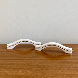 May include: Two white ceramic cabinet pulls with a gold accent line. The pulls are shaped like an arch.