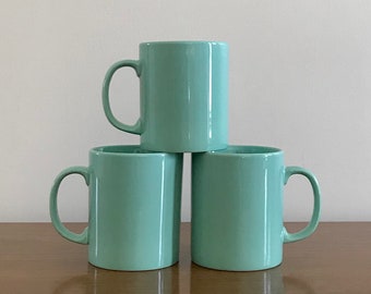 May include: Three light blue ceramic mugs stacked on top of each other on a wooden surface.